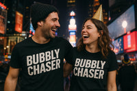 A man and woman smiling and laughing in a vibrant, possibly urban setting at night. Both are wearing matching black t-shirts with the text 
