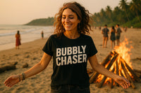 A person with curly hair is smiling and wearing a black t-shirt that says “BUBBLY CHASER.” They are standing on a beach near a bonfire, with other people and the ocean in the background. The atmosphere is warm and relaxed, suggestive of a sunset gathering by the sea.