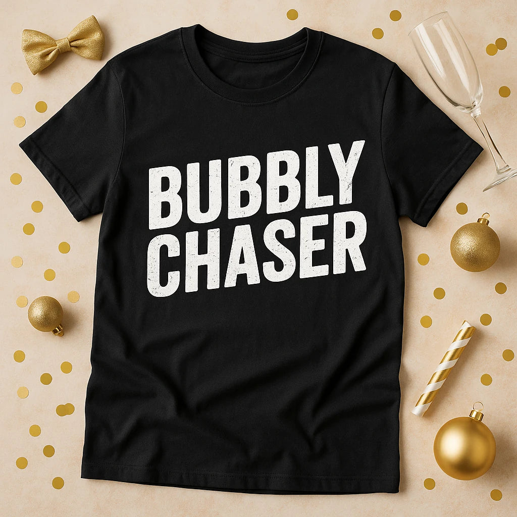 A black T-shirt with the words "BUBBLY CHASER" printed in large white letters. The shirt is laid flat on a tan background scattered with gold confetti, a gold bow, gold ornaments, a gold-striped straw, and a champagne flute placed nearby.