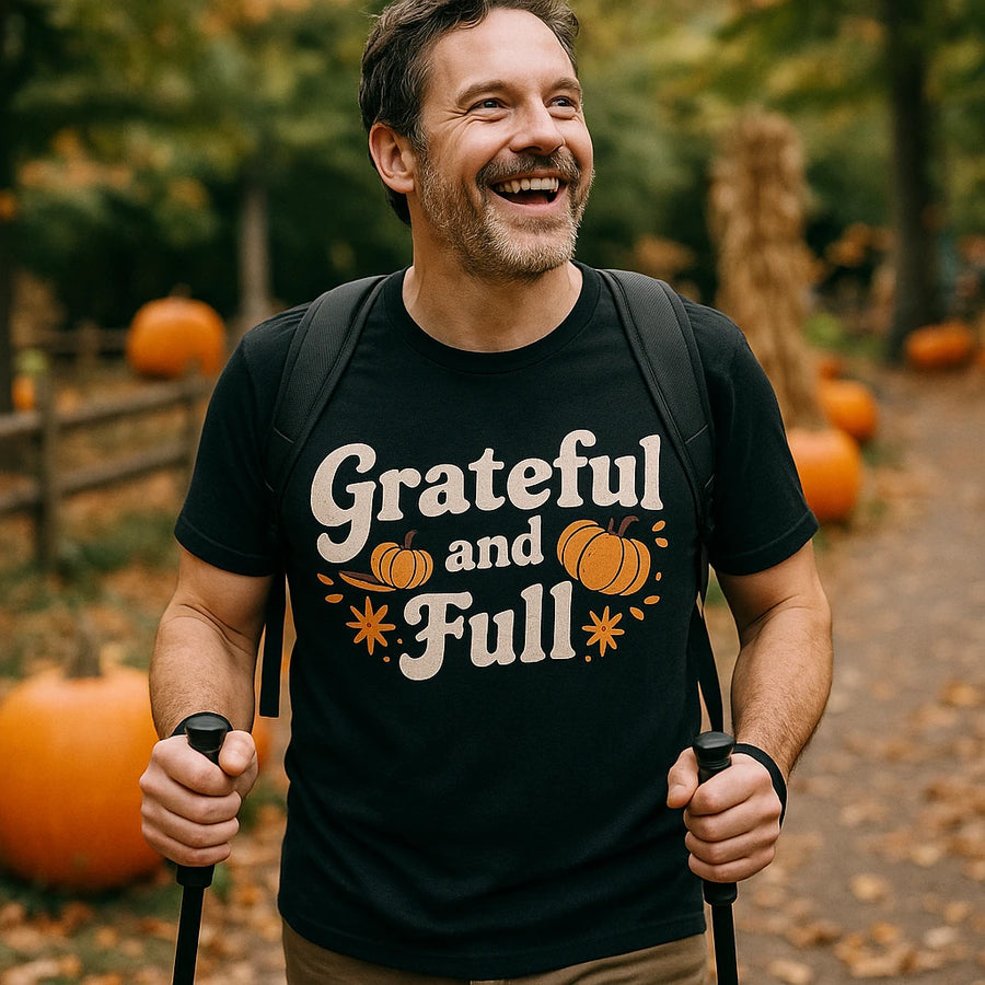 A man outdoors, smiling broadly while holding walking poles. He is wearing a black T-shirt with the phrase "Grateful and Full" and designs featuring pumpkins and stars. The background includes a fall scene with pumpkins and trees.
