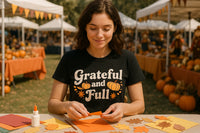 A person with brown hair is crafting paper leaves at a table outdoors, surrounded by a festive fall market setting with pumpkins and vendor stalls. The person is wearing a black t-shirt with the text 