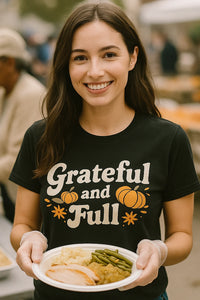 A person with long dark hair is smiling while holding a plate of food, which includes green beans, mashed potatoes, and slices of turkey. They are wearing a black T-shirt with text and pumpkin graphics. The background shows an outdoor setting with other people blurred in the distance.