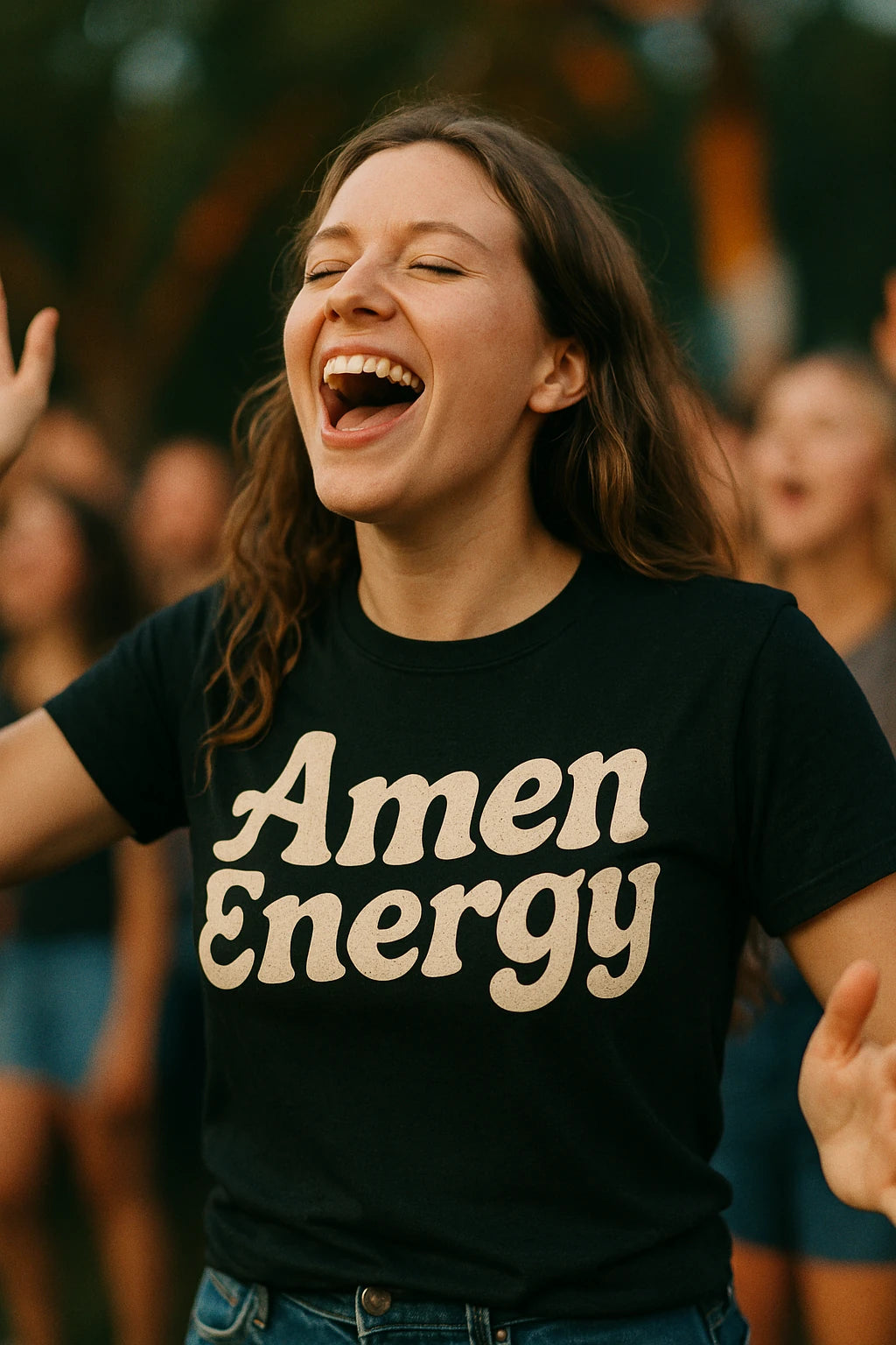A person with long brown hair is joyfully singing or shouting with their eyes closed and hands slightly raised. They are wearing a black T-shirt with the words "Amen Energy" printed in bold white letters. The background is blurry, suggesting an outdoor setting possibly at a gathering or event.