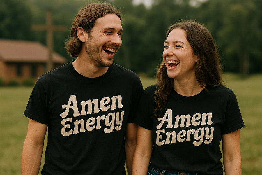 A man and a woman, both wearing black t-shirts with the text "Amen Energy," are standing outdoors and laughing. The background features greenery and a building with a cross in the distance.