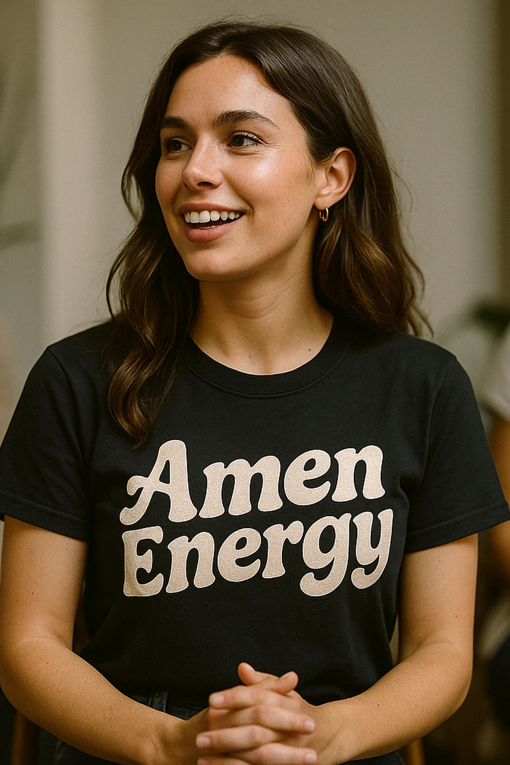 A person with long brown hair is smiling, wearing a black t-shirt with the text "Amen Energy." They have small hoop earrings and are standing with hands clasped.