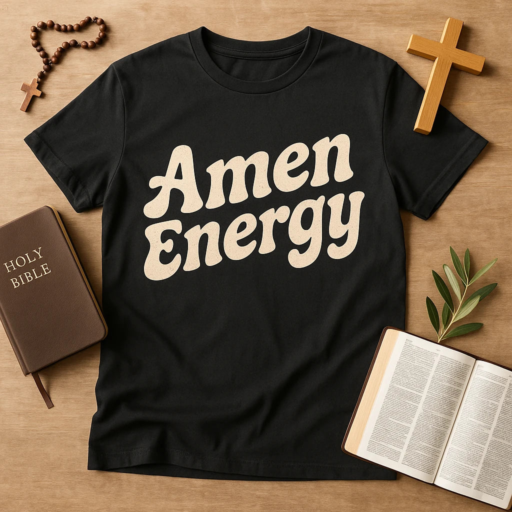 A black t-shirt with the text "Amen Energy" in bold, cream-colored letters. The shirt is laid out on a wooden surface surrounded by religious items: a closed Holy Bible with a brown cover, an open Bible with readable text, a wooden cross, and a rosary with a wooden cross. There are also a few green leaves placed near the open Bible.