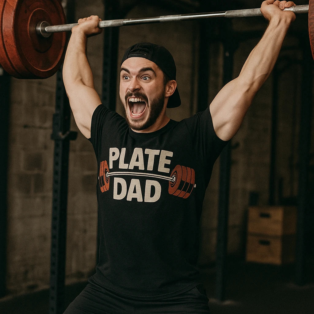 A person is lifting a barbell with red weight plates while wearing a black T-shirt with the text "PLATE DAD" and a graphic of a barbell. They are in a gym setting and appear to be expressing excitement or effort.