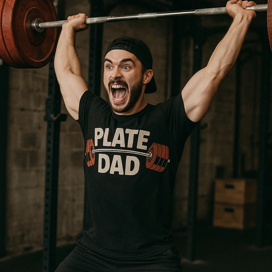 A person is lifting a barbell with red weight plates while wearing a black T-shirt with the text "PLATE DAD" and a graphic of a barbell. They are in a gym setting and appear to be expressing excitement or effort.
