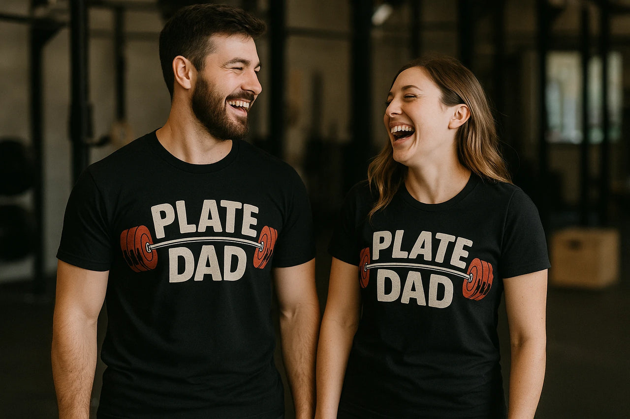 A man and woman stand next to each other, both wearing black t-shirts with the text "PLATE DAD" and an illustration of a barbell with red weights. They are inside what appears to be a gym and are both smiling at each other, exuding a joyful and friendly atmosphere.