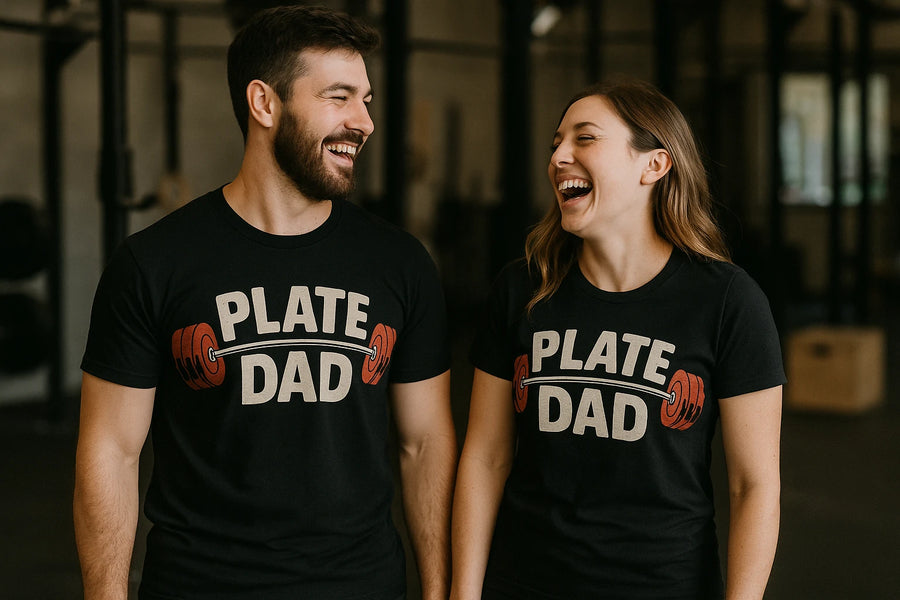 A man and woman stand next to each other, both wearing black t-shirts with the text "PLATE DAD" and an illustration of a barbell with red weights. They are inside what appears to be a gym and are both smiling at each other, exuding a joyful and friendly atmosphere.