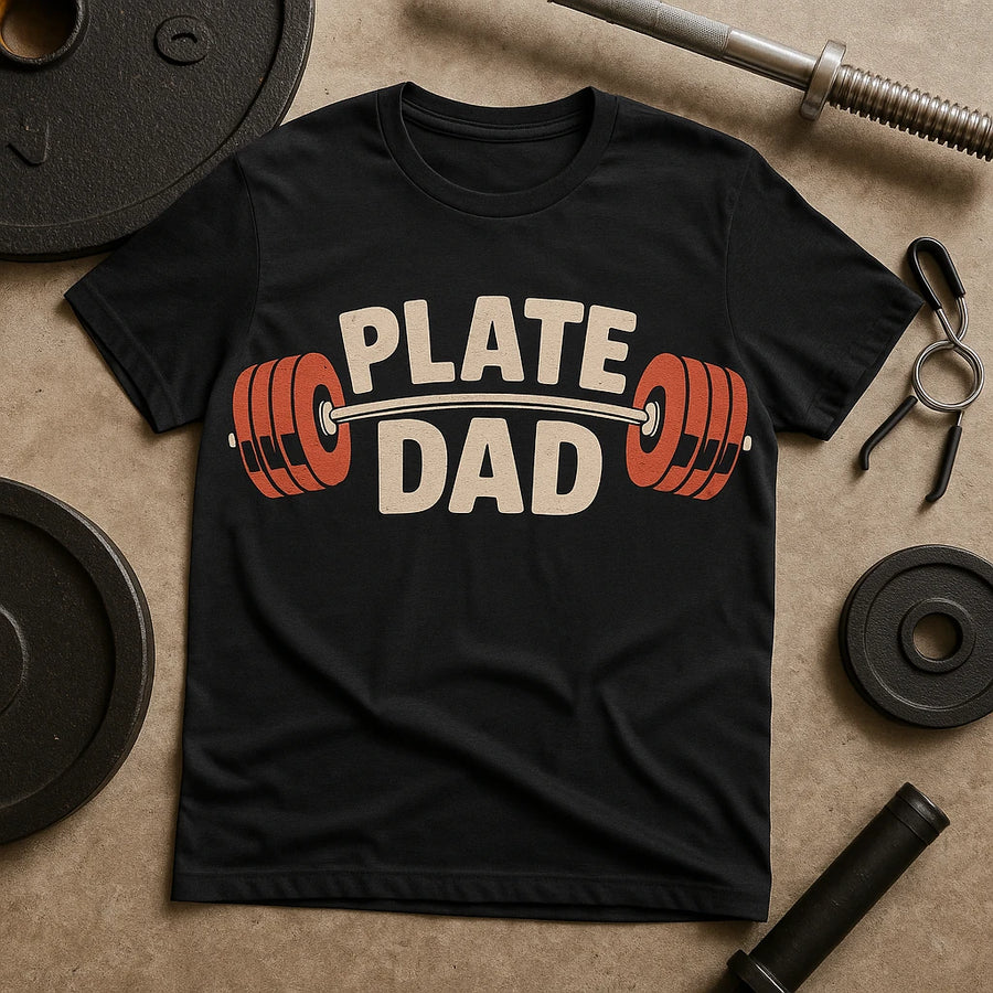 A black t-shirt with the text "PLATE DAD" written in bold letters. The text is adorned with a graphic of a barbell with weight plates on either side. The t-shirt is surrounded by gym equipment, including weight plates, a barbell, and weight collars, all placed on a textured surface.