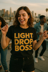 A person with shoulder-length brown hair is smiling and dancing outdoors with a city skyline in the background. They are wearing a black t-shirt with the text 