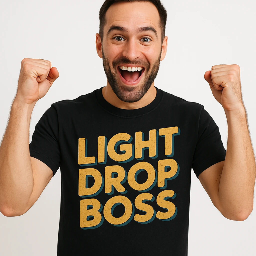 A person with short dark hair and a beard is wearing a black T-shirt with the text "LIGHT DROP BOSS" printed in bold, mustard-yellow letters. They are smiling broadly with fists raised in excitement, standing against a plain white background.