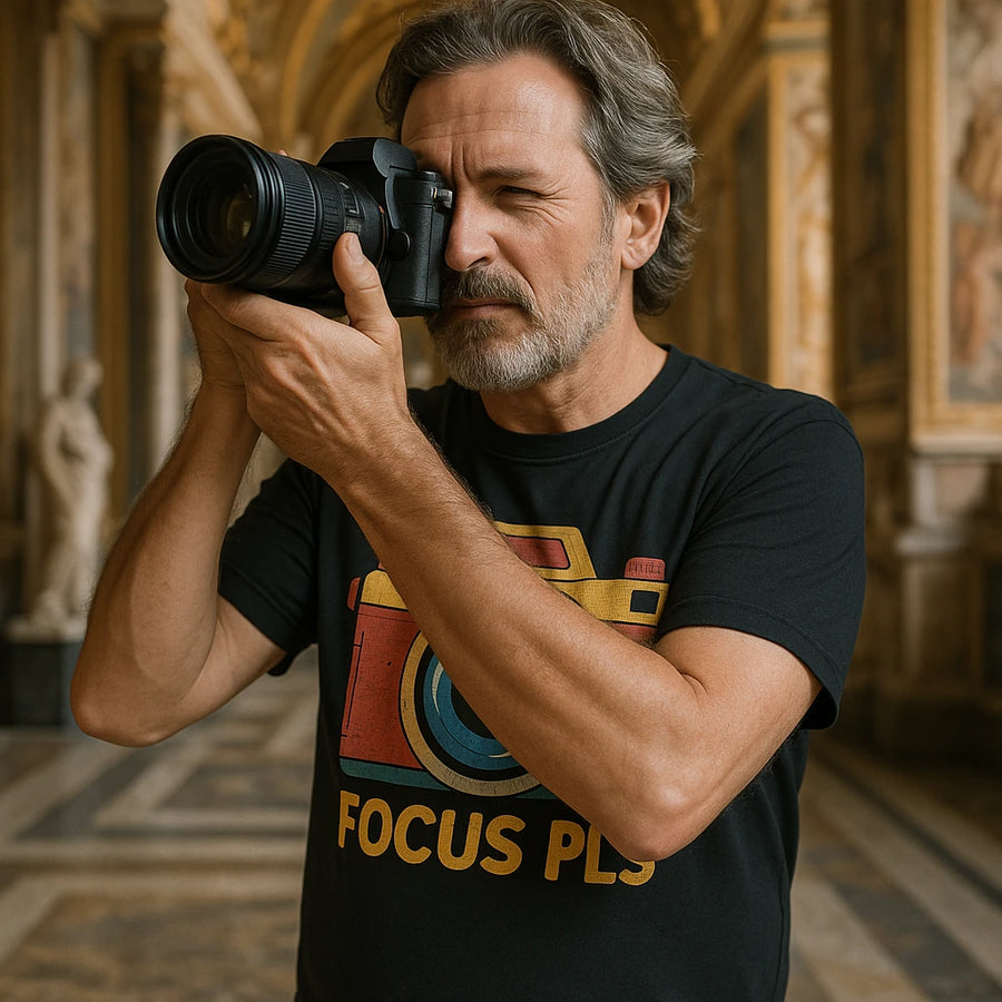 A bearded man with gray hair is wearing a black T-shirt with a colorful camera graphic and the text "FOCUS PLS." He is holding a camera up to his eye, appearing to take a photograph. The setting is an ornate interior with marble floors and classical sculptures in the background.