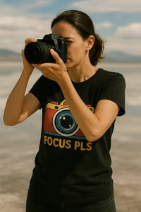 A person with dark hair is taking a photo with a DSLR camera. They are wearing a black t-shirt featuring a colorful camera graphic and the text 
