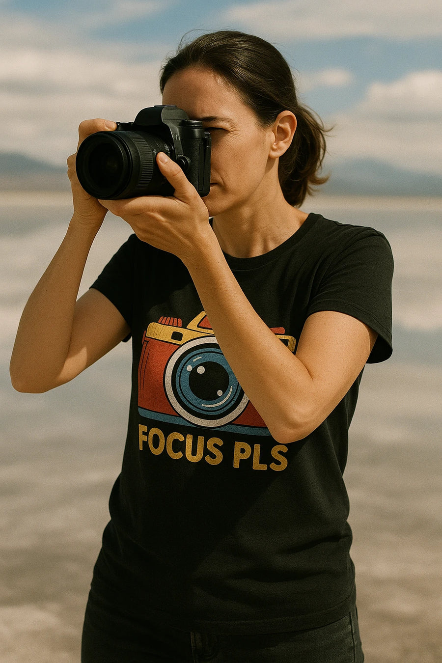 A person with dark hair is taking a photo with a DSLR camera. They are wearing a black t-shirt featuring a colorful camera graphic and the text "FOCUS PLS." The background appears to be an outdoor setting with a cloudy sky.