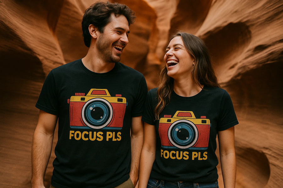 A man and woman are laughing and looking at each other, wearing matching black t-shirts with a colorful camera graphic and the words "FOCUS PLS" printed on them. The background appears to be a canyon with textured, tan rock formations.