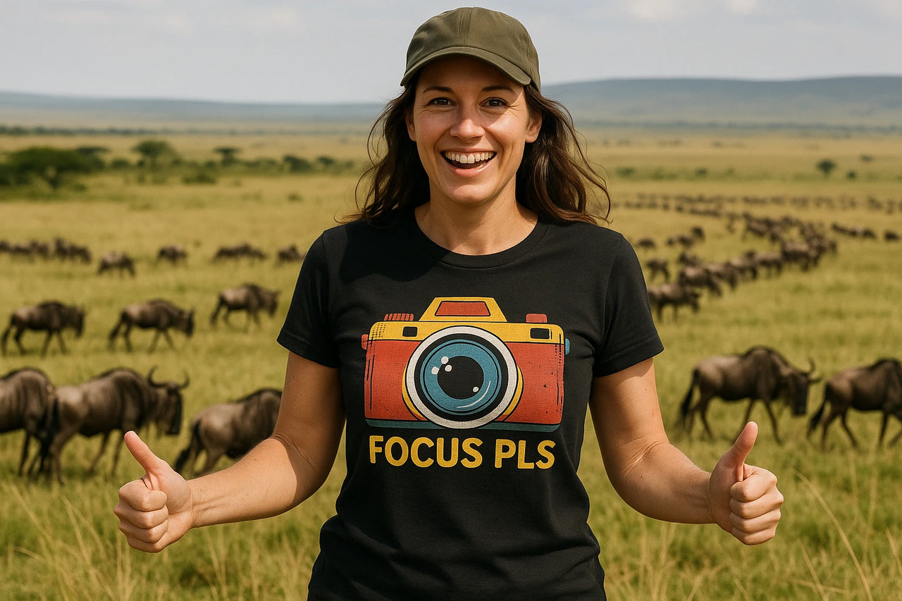 A person smiling and giving two thumbs up, wearing a black shirt with a colorful camera graphic and the text "FOCUS PLS." They have long hair and a green cap. In the background is a vast savannah with a herd of wildebeests.