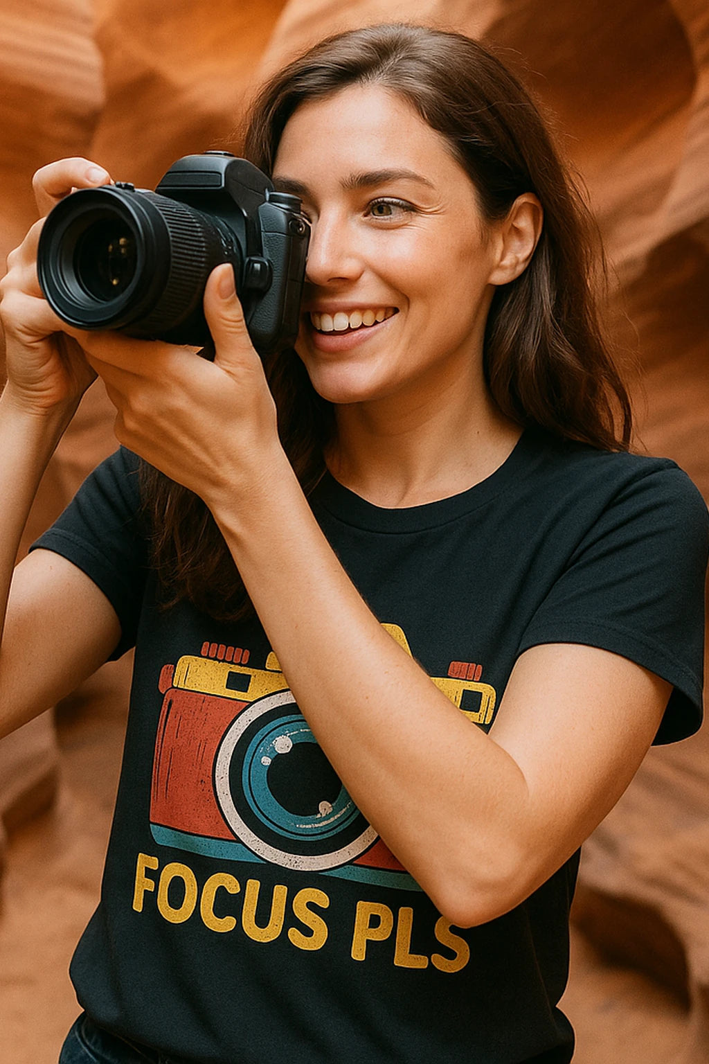 A person with long brown hair is smiling while holding a camera up to their eye. They are wearing a black t-shirt featuring a colorful camera graphic and the words "FOCUS PLS." The background appears to be a sandstone canyon.