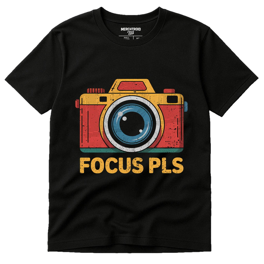 A black t-shirt with a colorful graphic of a camera above the text "FOCUS PLS" in bold yellow letters. The design features a stylized illustration with red, yellow, and blue elements, giving a retro vibe.