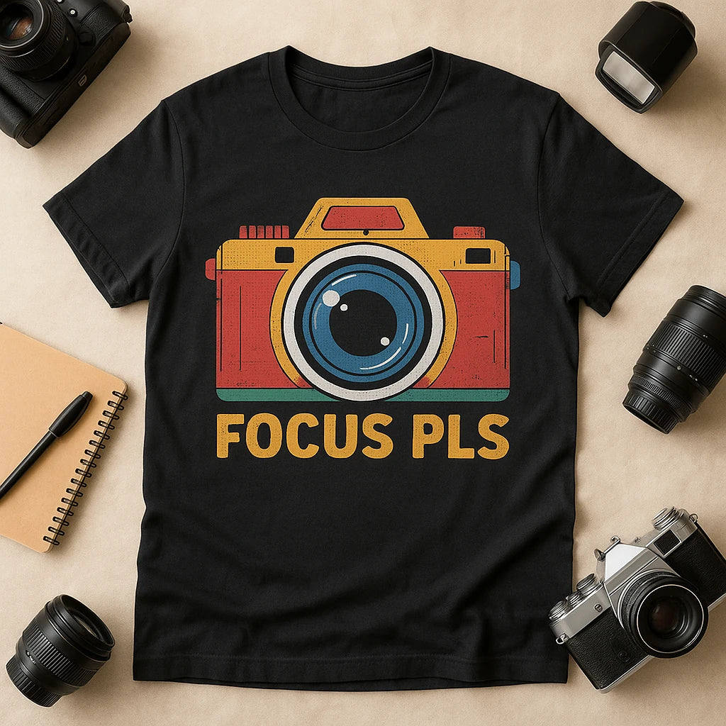 A black t-shirt featuring a colorful illustration of a camera with the text "FOCUS PLS" below it. The shirt is laid flat surrounded by camera equipment, including lenses and a notebook with a pen.