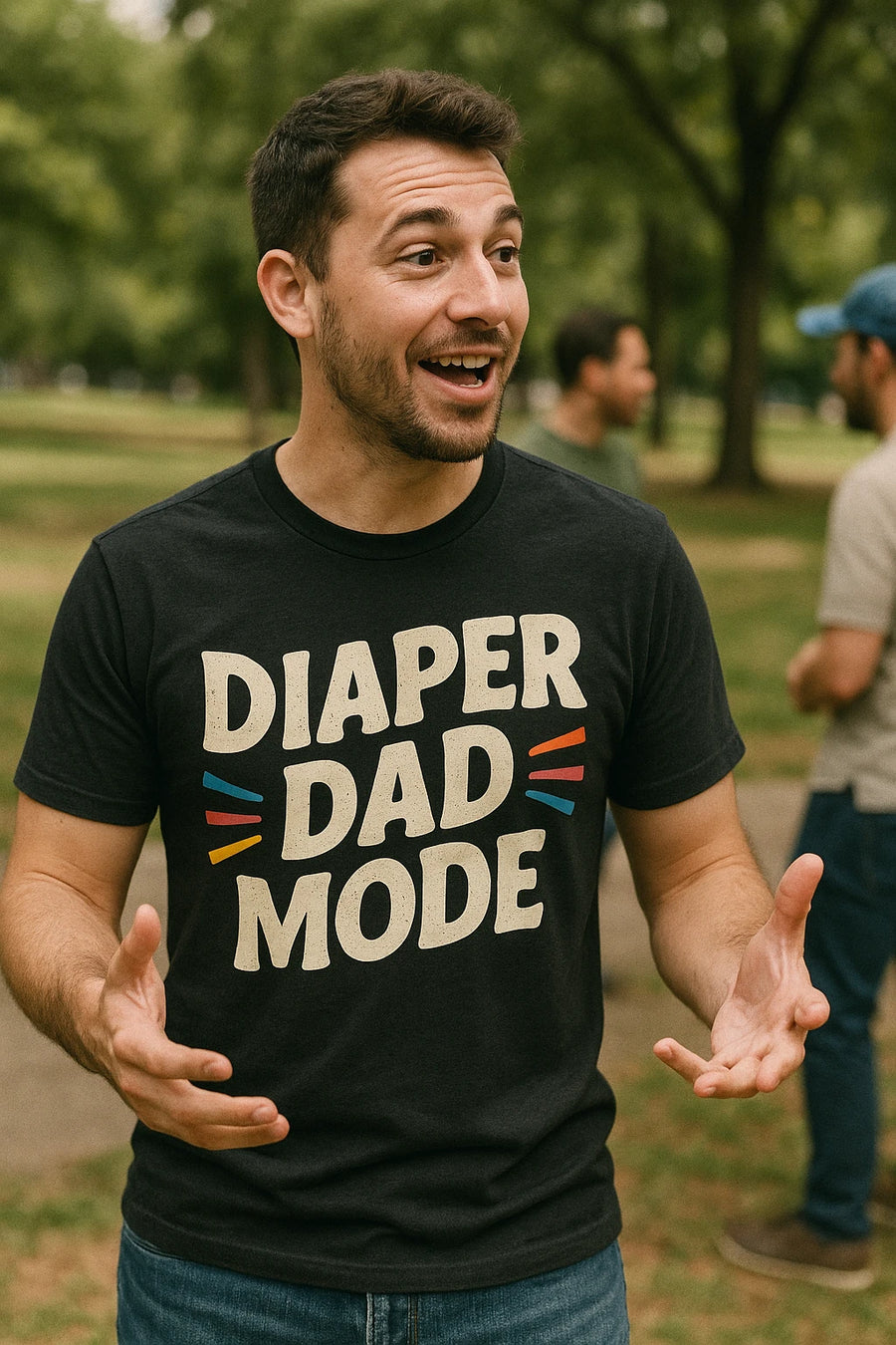 A person with short dark hair and a beard is wearing a black t-shirt that reads "DIAPER DAD MODE" in bold, white letters. They are standing outdoors, with blurred greenery in the background, gesturing with their hands and smiling.