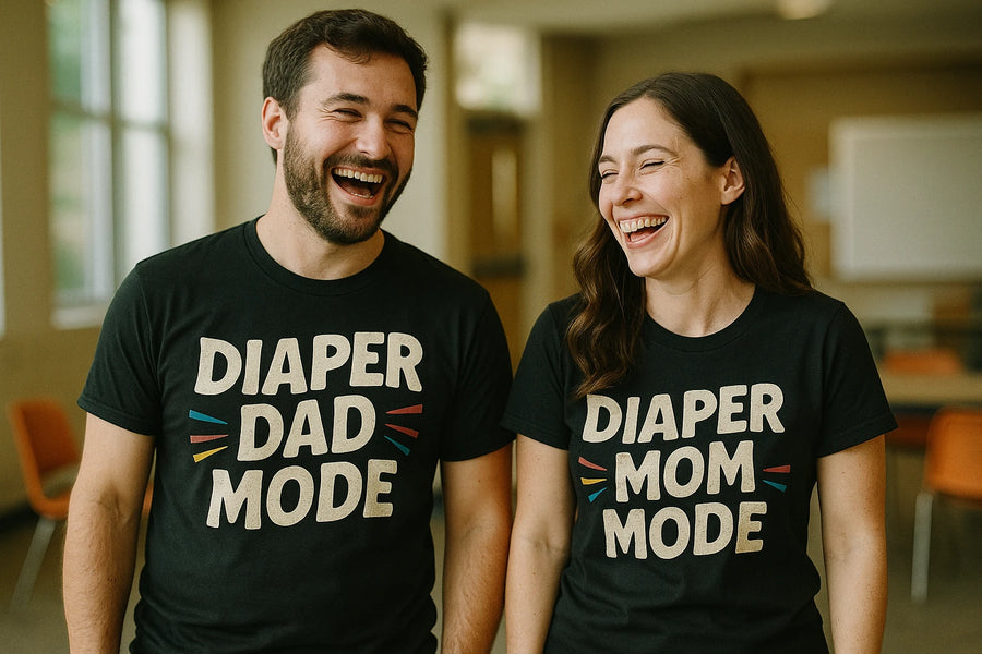A man and a woman are standing side by side, both laughing. The man has short dark hair and a beard, and the woman has long brown hair. They are wearing matching black T-shirts with humorous text. The background appears to be an indoor setting with orange chairs and natural light coming through the windows.