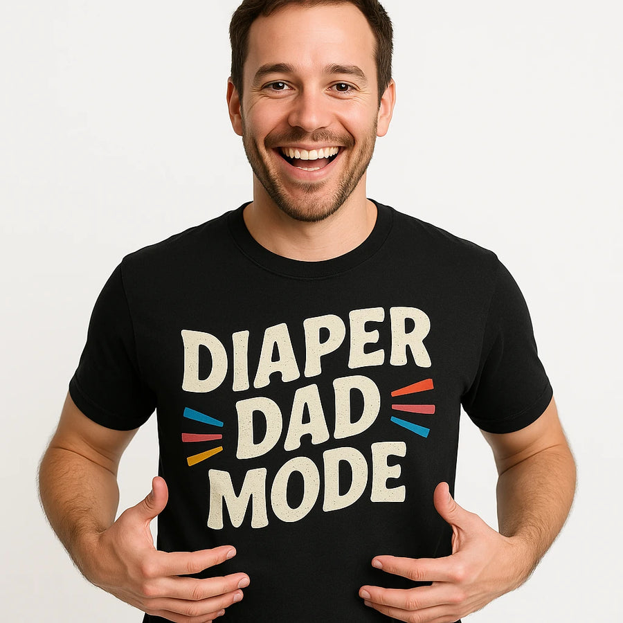 A smiling man with short dark hair and a beard is wearing a black T-shirt with the text "DIAPER DAD MODE" on it. The text is in a large, bold, white font with colorful stripes around "DAD". The background is plain white.