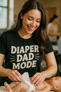 A person with long brown hair is smiling while changing a diaper on a baby doll. They are wearing a black T-shirt with the text 