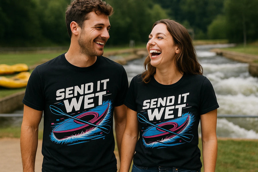 Two people are outdoors near a flowing waterway, both smiling and looking at each other. They are wearing matching black t-shirts with the text "SEND IT WET" and graphic art of a kayak and paddle. There is greenery and a water feature in the background.