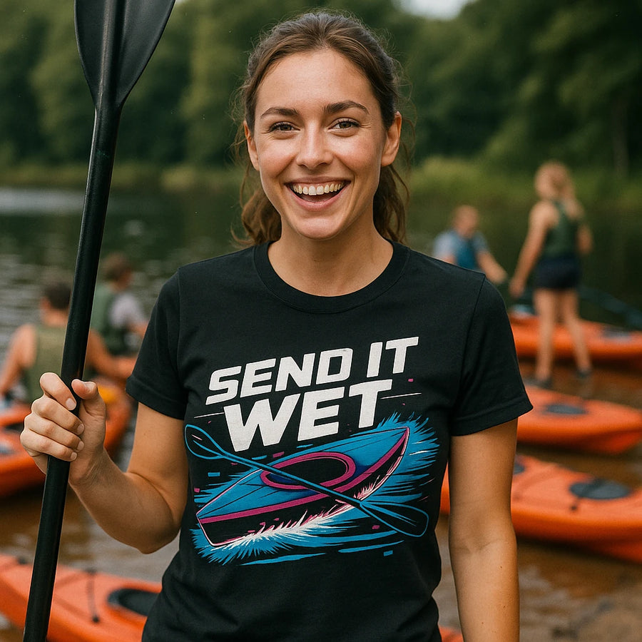 A person is smiling and holding a paddle, standing in front of kayaks on a lake. They are wearing a black t-shirt with a colorful graphic of a kayak and the text "SEND IT WET." The background shows several people near the water with more kayaks. The setting appears to be outdoors in a natural area with trees.