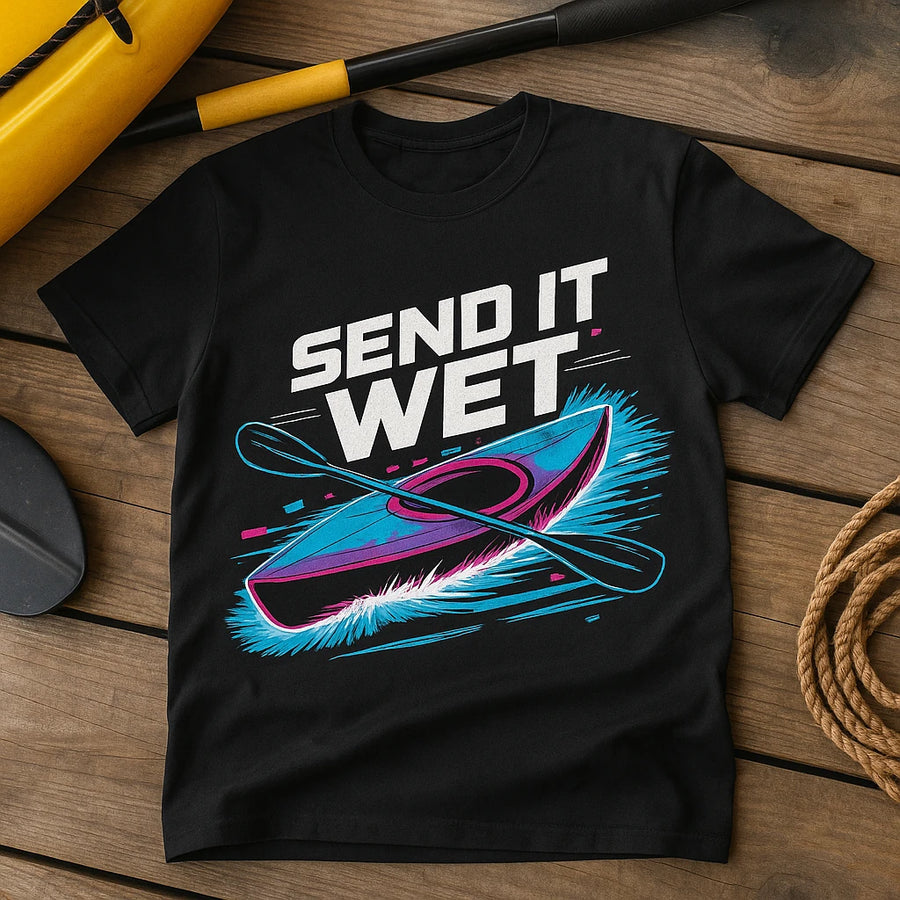 A black t-shirt is laid flat on a wooden surface, featuring a graphic of a kayak and paddle with the text "SEND IT WET" in bold, white letters. The design includes vibrant blue and pink accents. Nearby are a yellow paddle and coiled rope.