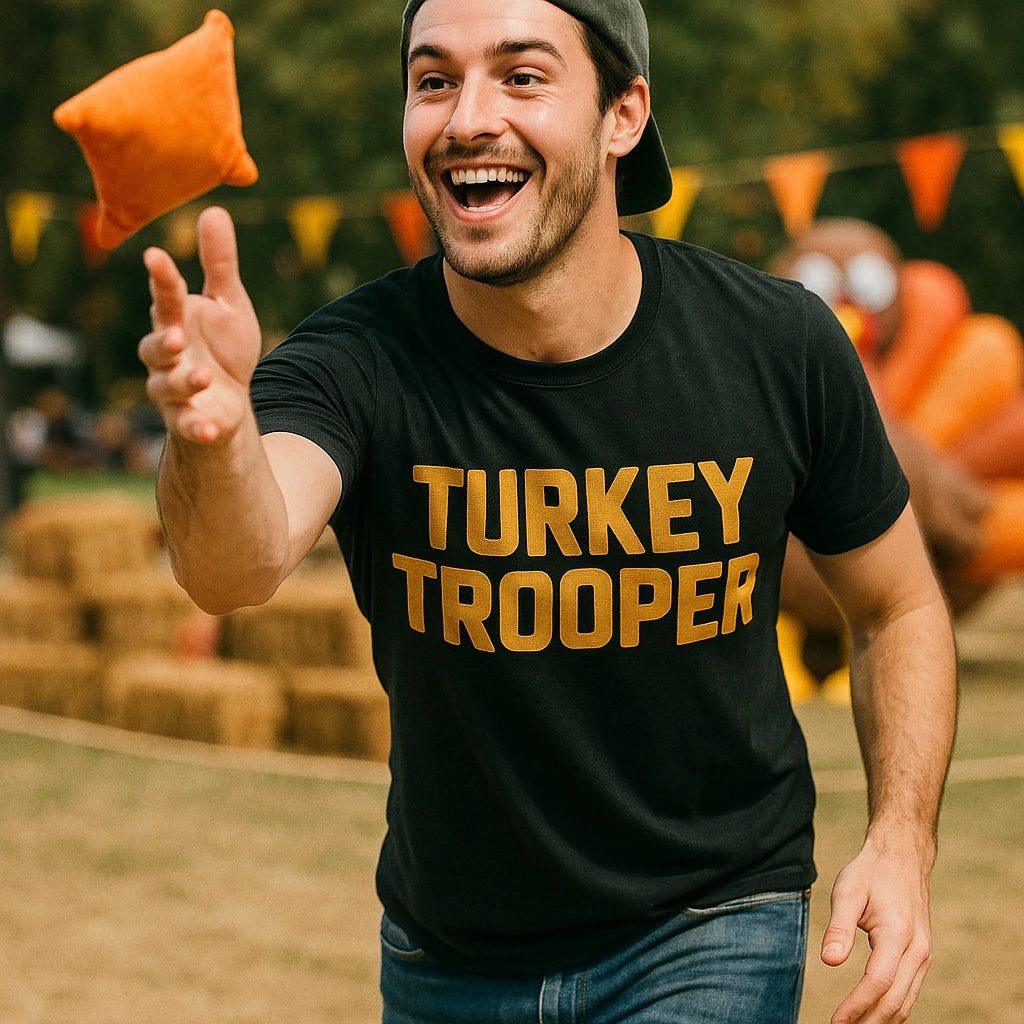 A person wearing a black T-shirt with the words "TURKEY TROOPER" in bold, playing an outdoor game involving an orange bean bag. The person appears to be smiling and enjoying the activity, with a blurred background featuring hay bales and colorful triangular flags.