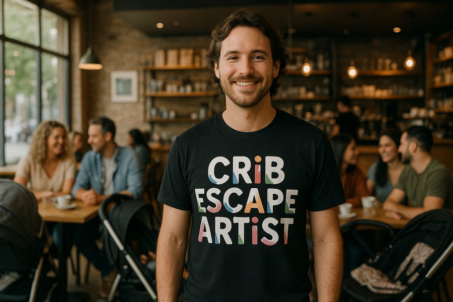A person is smiling and standing in a cozy café environment. They are wearing a black t-shirt with the text "CRiB ESCAPE ARTiST" in colorful letters. In the background, there are several people sitting at tables, engaged in conversation, with soft lighting enhancing the warm atmosphere.