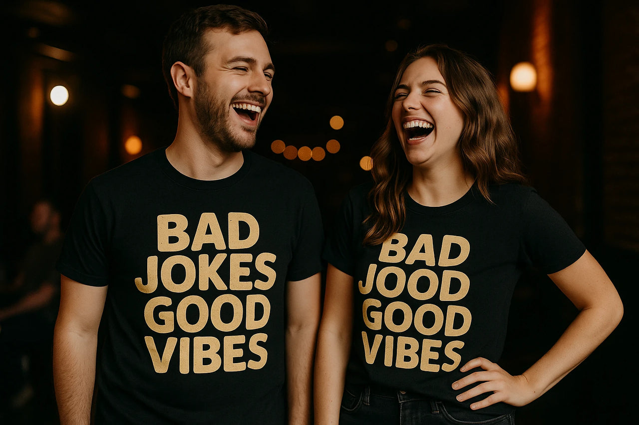 Two people standing side by side, both wearing black t-shirts with gold text. The person on the left has short hair and facial hair, while the person on the right has long, wavy hair. Both are laughing joyfully in a dimly lit indoor setting with warm lighting in the background.