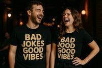 Two people standing side by side, both wearing black t-shirts with gold text. The person on the left has short hair and facial hair, while the person on the right has long, wavy hair. Both are laughing joyfully in a dimly lit indoor setting with warm lighting in the background.