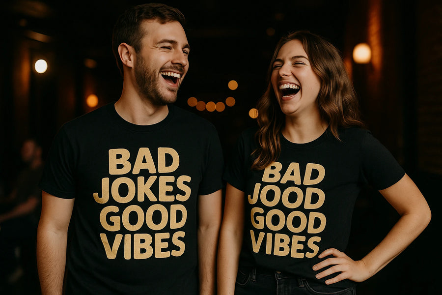 Two people standing side by side, both wearing black t-shirts with gold text. The person on the left has short hair and facial hair, while the person on the right has long, wavy hair. Both are laughing joyfully in a dimly lit indoor setting with warm lighting in the background.