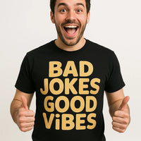 A person with a beard and short dark hair, smiling widely and giving two thumbs up. They are wearing a black t-shirt with bold yellow text. The background is plain white.