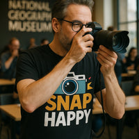 A person with glasses is holding a camera up to their face as if taking a photo. They are wearing a black t-shirt with a colorful design of a camera and the words 