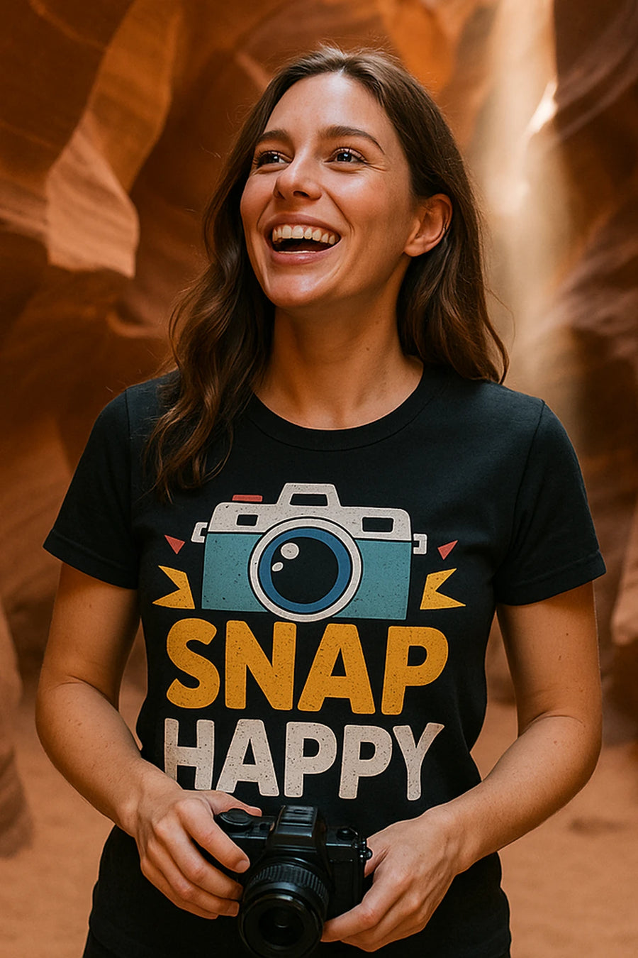 A person with long hair is smiling and holding a camera in both hands. They are wearing a black T-shirt with a graphic of a camera and the words "SNAP HAPPY." The background features red rock formations and beams of light.
