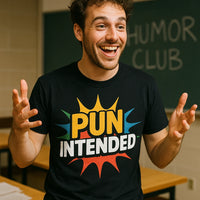 A person with curly hair is standing in a classroom, smiling and gesturing with both hands. They are wearing a black T-shirt with colorful text that reads 