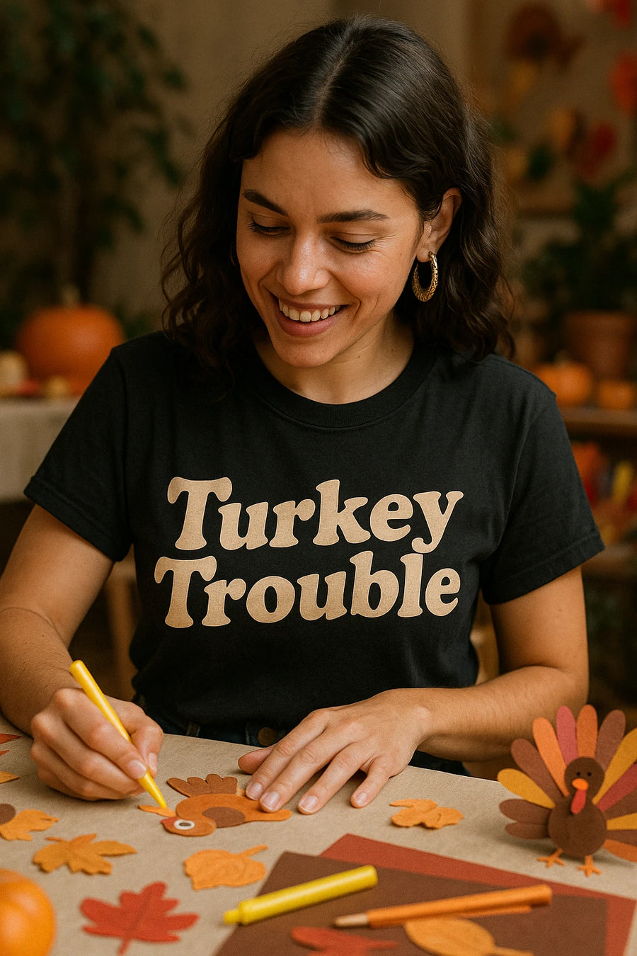 A person with curly dark hair and wearing gold hoop earrings is smiling while engaged in a craft activity. They have a black T-shirt with the text "Turkey Trouble" in bold letters. The table is covered with autumn-themed crafts, including leaves and a paper turkey. Pumpkins are visible in the background.