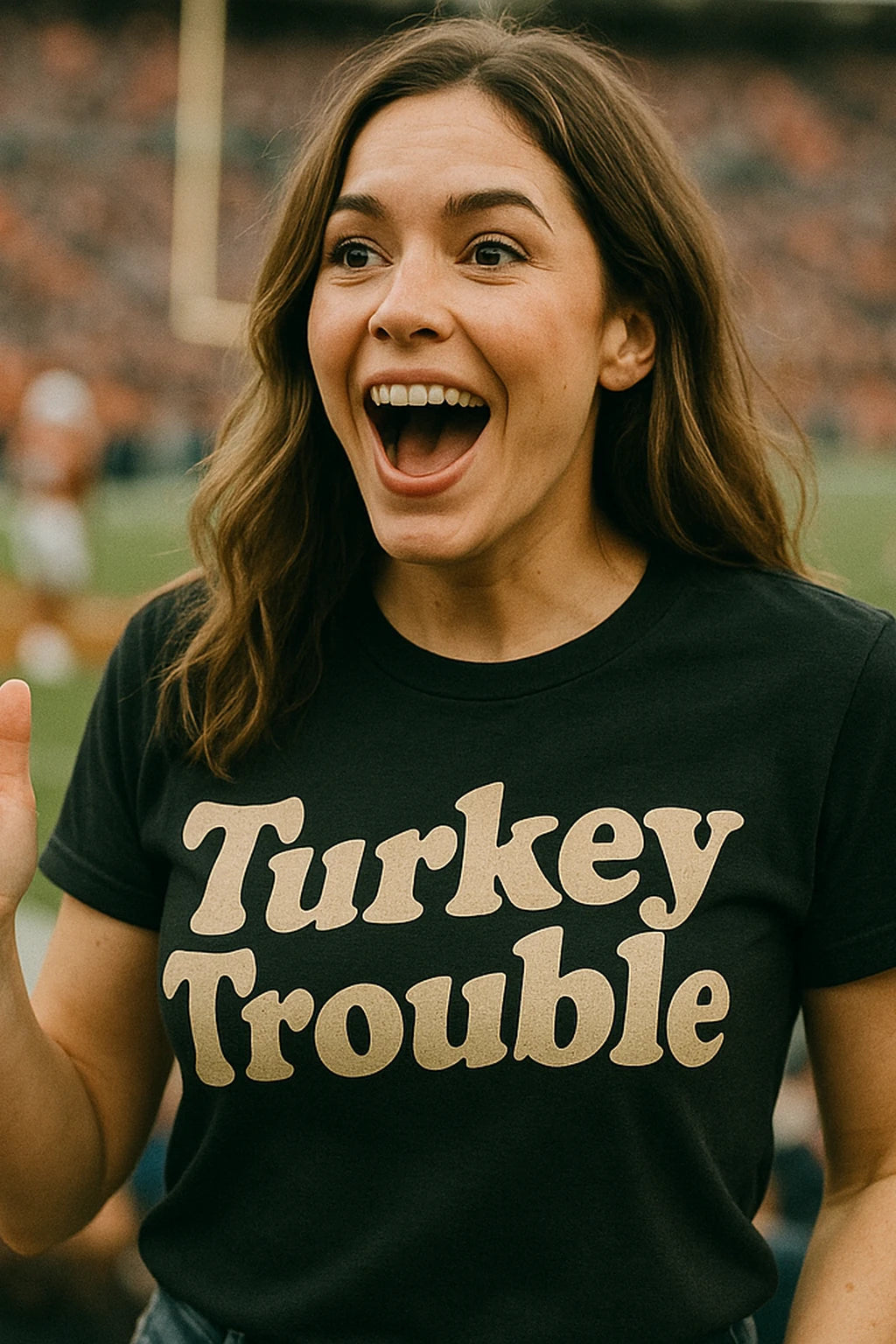 A person with long brown hair is standing in a stadium, looking excited and smiling widely. They are wearing a black t-shirt with the text "Turkey Trouble" printed in bold letters. The background is blurred, showing a sports field with a goalpost.