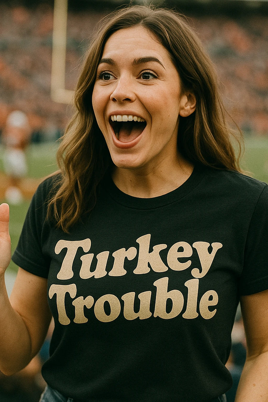 A person with long brown hair is standing in a stadium, looking excited and smiling widely. They are wearing a black t-shirt with the text "Turkey Trouble" printed in bold letters. The background is blurred, showing a sports field with a goalpost.
