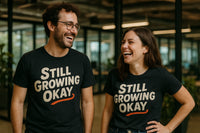 A man and a woman are standing indoors, both smiling and wearing black t-shirts with the words 