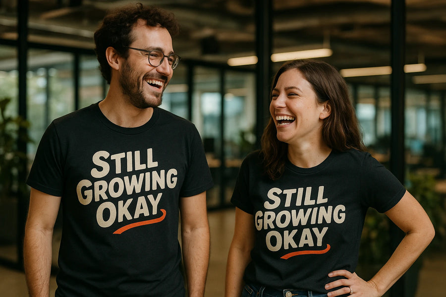 A man and a woman are standing indoors, both smiling and wearing black t-shirts with the words "STILL GROWING OKAY" printed on them. The background shows a modern office environment with glass walls and soft lighting.