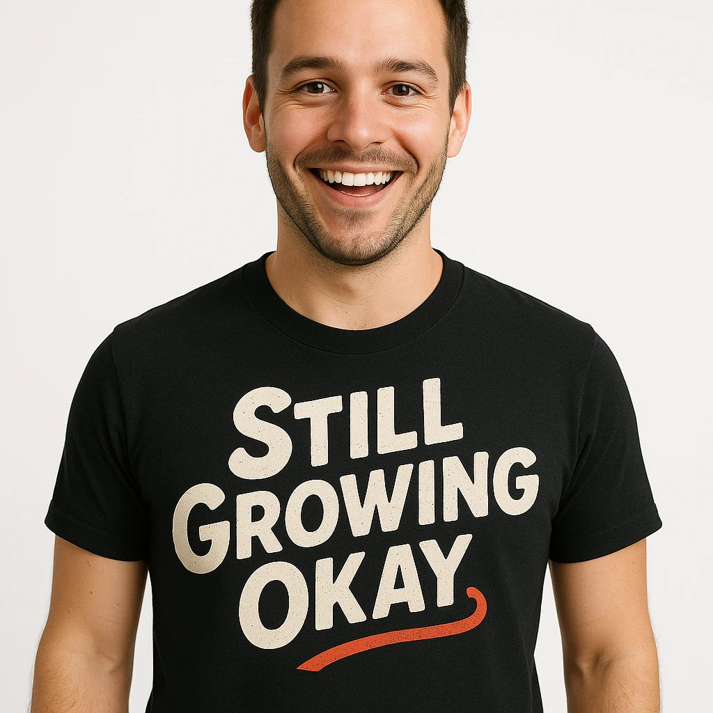 A person with short dark hair and a beard is smiling, wearing a black T-shirt with the text "STILL GROWING OKAY" in bold white letters and a red underline. The background is plain white.