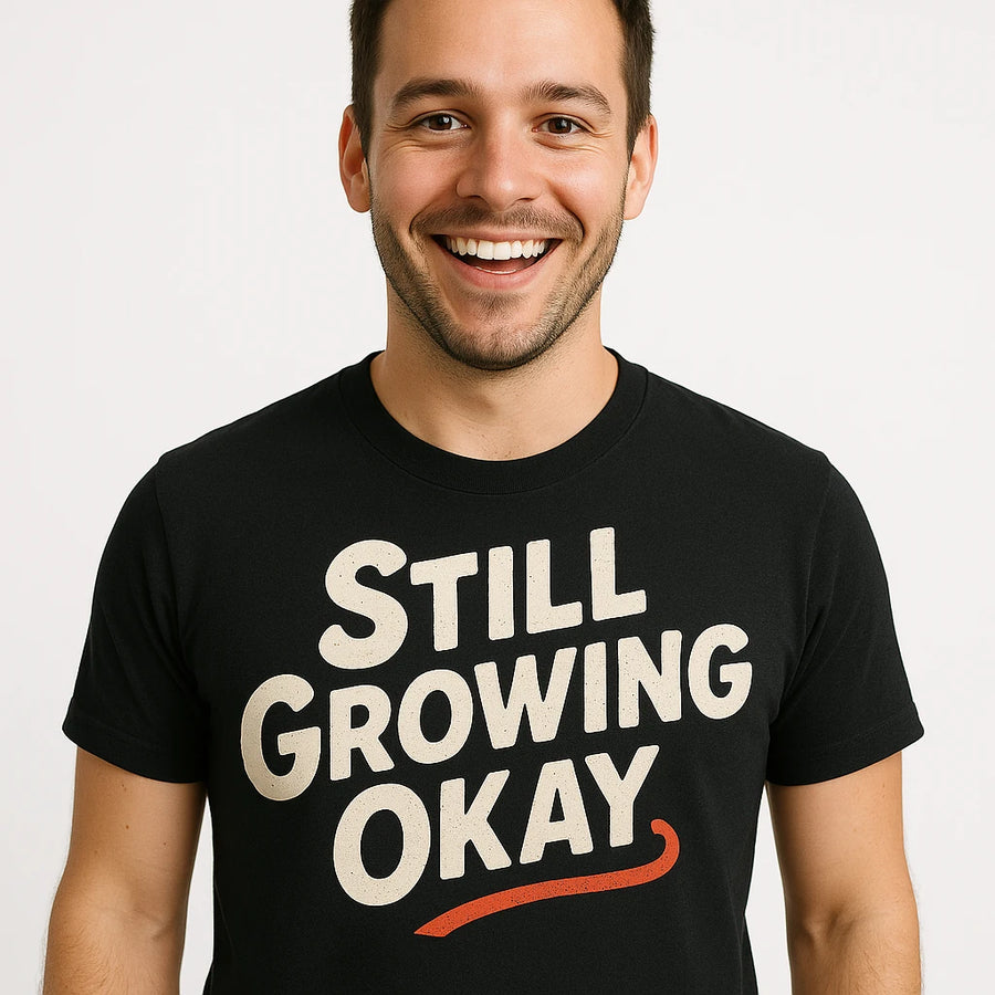 A person with short dark hair and a beard is smiling, wearing a black T-shirt with the text "STILL GROWING OKAY" in bold white letters and a red underline. The background is plain white.