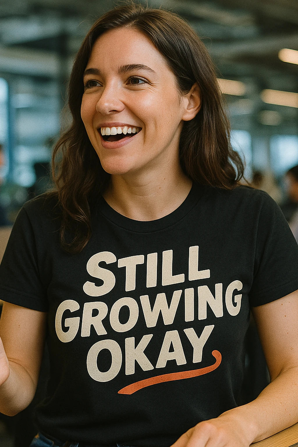 A person with long brown hair is smiling and wearing a black shirt with the text "STILL GROWING OKAY." The background is a blurred indoor setting.