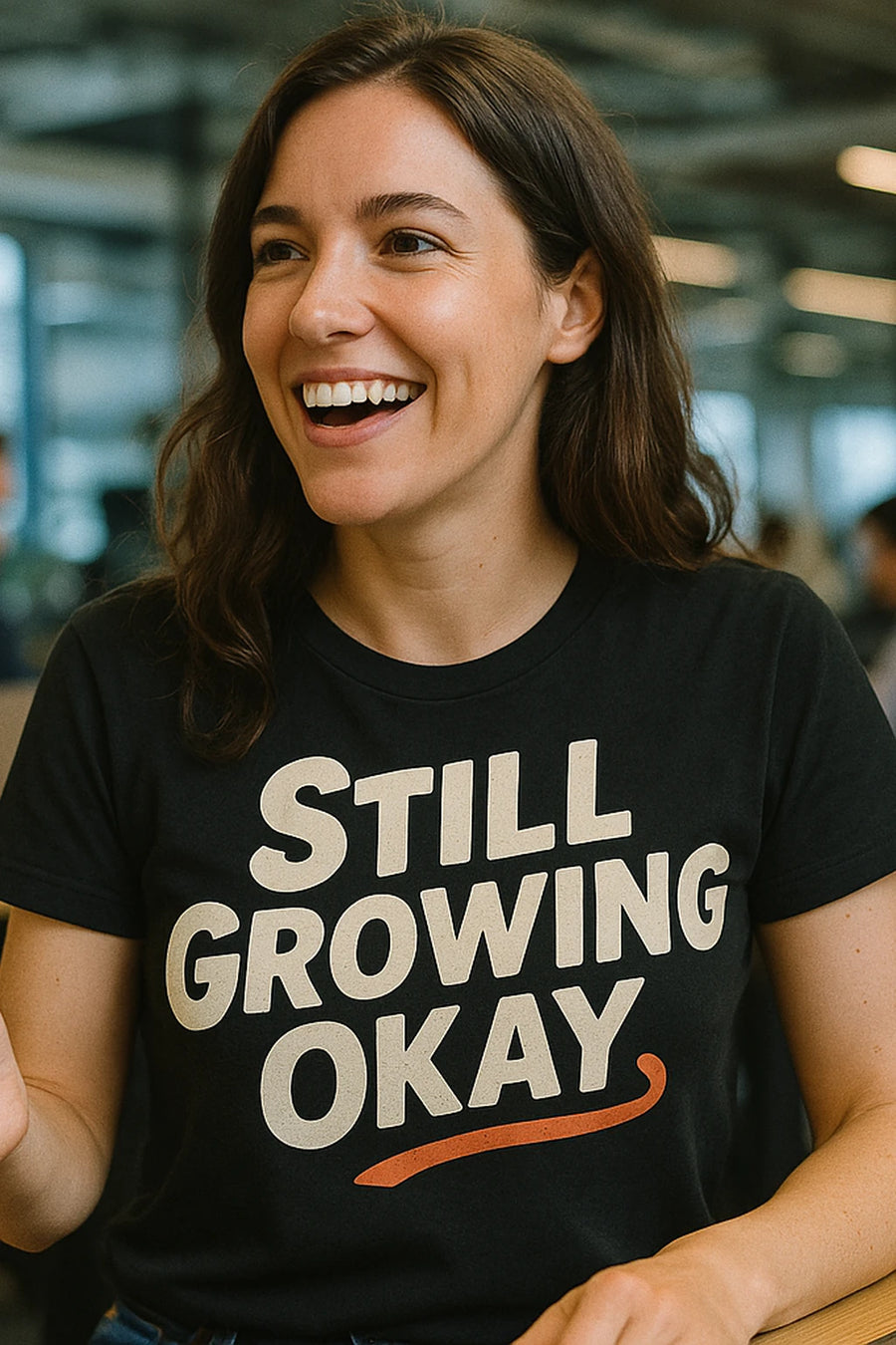 A person with long brown hair is smiling and wearing a black shirt with the text "STILL GROWING OKAY." The background is a blurred indoor setting.